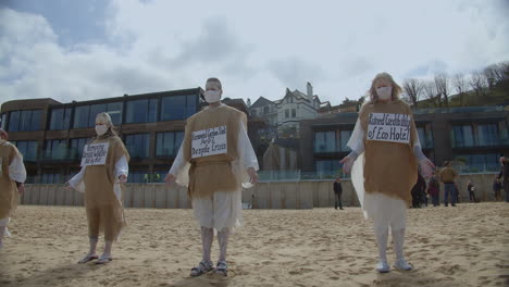 A-number-of-protesters-stand-motionless-on-a-beach-in-front-of-the-Carbis-Bay-Hotel-in-St-Ives,-Cornwall,-UK