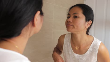 Young-asian-woman-standing-in-front-of-a-mirror-looking-at-the-reflection-checking-acne-on-her-face-with-a-converned-look-on-her-face