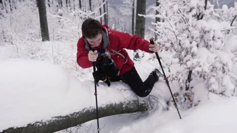 Young-male-hiker-with-hiking-poles-climbing-over-snowy-wooden-trunk-in-forest
