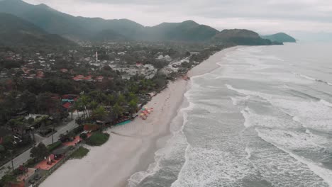 Lateral-drone-footage-of-the-beach,-mountain-and-houses-in-front-of-the-sea,-cloudy-day,-waves,-landscape-of-Juquehy,-Ubatuba,-northern-coast-of-São-Paulo,-Brazil