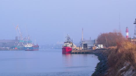 Large-blue-cargo-ship-and-fishing-vessels-at-Port-of-Liepaja-in-foggy-day,-calm-day-with-mist,-distant-medium-shot