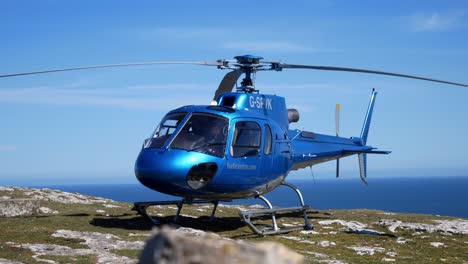 Private-tour-helicopter-on-mountain-cliff-summit-overlooking-blue-ocean-landscape-take-off-preparation