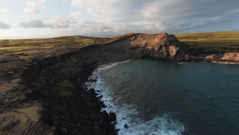 Fpv-aerial-shot-showing-crashing-waves-of-the-ocean-against-rocky-cliff-wall-and-green-lighting-landscape-in-background