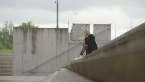 Young-blond-woman-sitting-outdoors-at-stairs-and-checking-smartphone-during-daytime