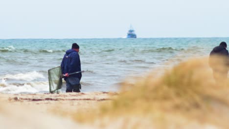 Two-men-with-fishnet-searching-natural-amber-stones-at-Baltic-sea-coastline-in-overcast-spring-day,-fishing-boat-in-background,-heat-waves,-medium-shot-from-a-distance