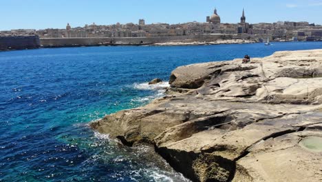 Aerial-view-towards-Valletta,-Malta-with-waves-splashing-rocky-limestone-Sliema-beach-in-sunny-summer-day