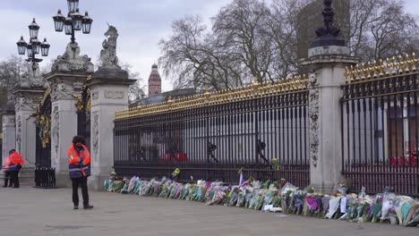 Buckingham-Palace-attendant-walks-in-front-of-the-gates-with-flowers-and-tributes,-after-the-death-of-Prince-Philip,-Duke-of-Edinburgh,-Saturday-April-10th,-2021---London-UK