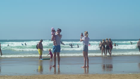Familia-Feliz-Tomando-Fotos-En-La-Playa-Con-Bañistas-Divirtiéndose-En-El-Fondo
