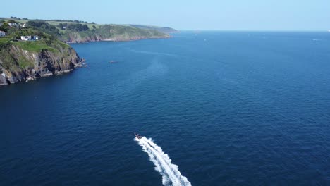 A-following-drone-shot-of-a-fast-rigid-inflatable-boat-that-powers-off-to-the-right,-whilst-the-camera-tilts-up-revealing-a-beautiful-coastal-scene-with-stunning-houses-perched-on-the-cliffs