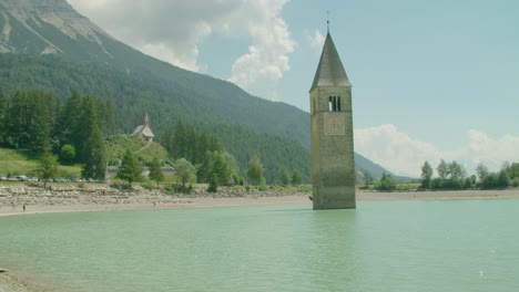 Full-shot,-Scenic-view-of-People-walking-on-the-shore-of-Reschensee-on-a-bright-sunny-day-in-Italy,-Kirchturm-von-Altgraun-in-the-background