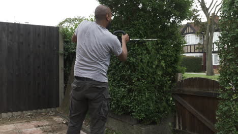 UK-Asian-Adult-Bald-Male-Using-Electric-Hedge-Trimmer-Outside-In-Front-Garden
