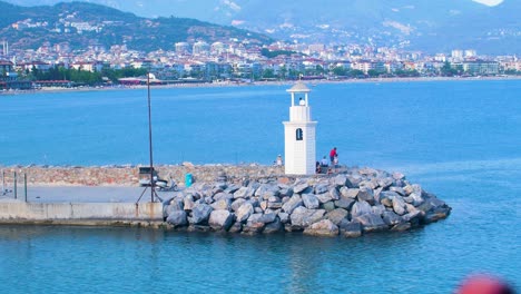 People-fishing-from-stone-pier-at-seaport-in-Alanya-,-small-white-lighthouse,-sunny-summer-day,-handheld-medium-shot