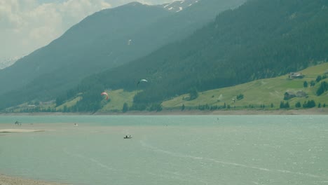 Volle-Aufnahme,-Malerische-Ansicht-Von-Menschen,-Die-Mitten-Im-Reschensee-In-Italien-Kitesurfen,-Ein-Heller-Sonniger-Tag-Im-Hintergrund
