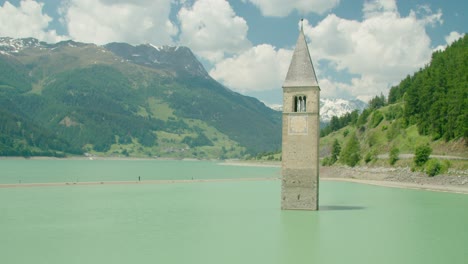 Volle-Aufnahme,-Malerischer-Blick-Auf-Den-Kirchturm-Von-Altgraun-Am-Reschensee-In-Italien,-Menschen,-Die-Auf-Dem-Seegrund-Spazieren-Gehen