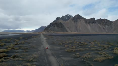 Drone-video-of-Stokksnes-Vestrahorn-black-sand-beach-in-Iceland-with-Red-car-driving-down-dirt-road