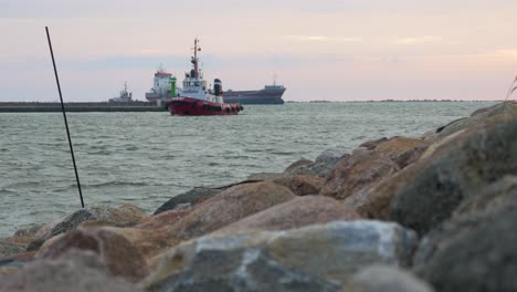 Red-and-white-port-pilot-ship-entering-the-Port-of-Liepaja-in-calm-sunny-summer-evening,-large-gray-cargo-ship-in-background,-stone-pier-in-foreground,-waves-splashing,-wide-low-angle-shot