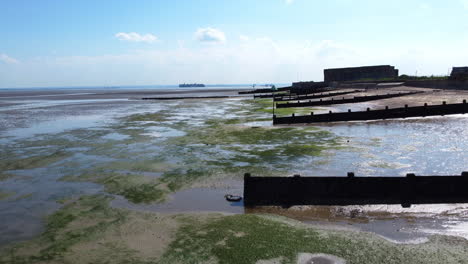 A-rising-drone-shot-of-an-abandoned-military-garrison-situated-behind-a-seawall-on-the-edge-of-beach-and-mudflats-on-a-tidal-estuary-at-low-tide,-on-a-clear-and-hot-summer-day