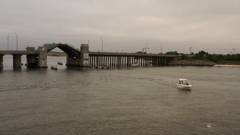 A-low-angle,-aerial-view-of-a-small-boat-sailing-on-Baldwin-Bay,-past-a-draw-bridge-opening-up,-on-a-cloudy-day