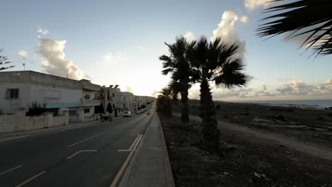 Malta,-Pembroke,-stunning-sunset-over-an-outer-road-and-sidewalk-with-palm-trees-and-commuting-cars-and-people