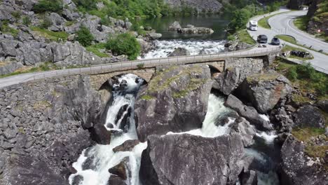Straße,-Die-Alte-Brücke-überquert,-Die-Von-Felsen-Gebaut-Wurde---Fluss,-Der-Vom-See-Und-Unter-Der-Brücke-Fließt---Mabodalen-Tal-Norwegen-Antenne