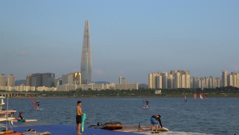 Korean-People-preparing-rig-and-sail-for-Windsurfing-on-Han-river-next-to-Lotte-World-Tower,-Seoul-on-Sunset