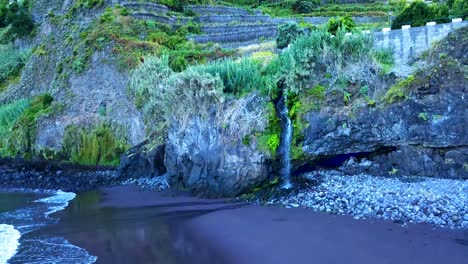 Pequeña-Cascada-Que-Fluye-Hacia-Las-Rocas-Y-La-Playa-De-Arena-Negra-En-Seixal,-Isla-De-Madeira,-Portugal