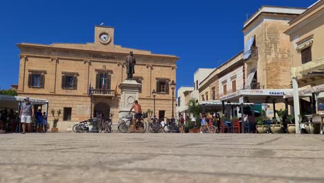 Low-angle-of-man-in-bicycle-at-Piazza-Europa-square-on-Favignana-island-with-Town-Hall-in-background,-Sicily