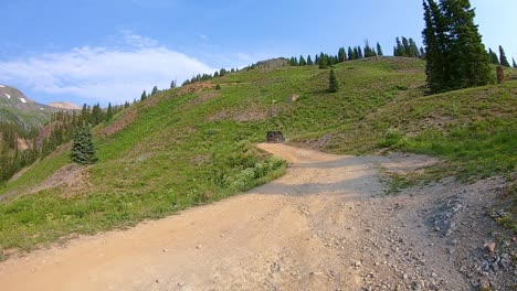 POV-following-a-Jeep-driving-up-Alpine-Loop-trail-cut-into-hillside,-through-meadow-in-San-Jan-Mountains-near-Silverton-Colorado