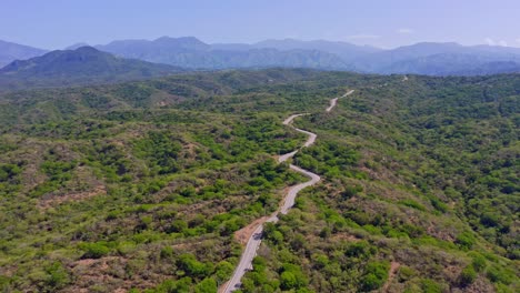 Cinematic-drone-flight-over-road-surrounded-by-green-planted-landscape-and-mountain-range-in-background---Hilly-countryside-in-San-Juan,Dominican-Republic