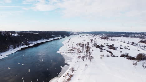 Flowing-winter-river-and-landscape-during-snowfall,-aerial-drone-view