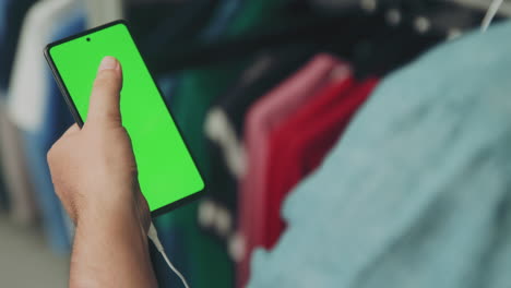 Mann-Im-Hemd-Mit-Handy-Mit-Grünem-Bildschirm,-Der-Durch-Das-Display-Scrollt,-Während-Er-In-Einem-Bekleidungsgeschäft-über-Die-Kopfhörer-Zuschaut-Und-Zuhört
