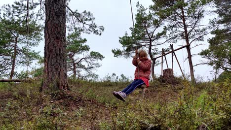 Young-girl-in-pink-riding-natural-swing-made-of-rope-and-a-branch---Having-fun-outdoors-alone-in-nordic-pine-forest