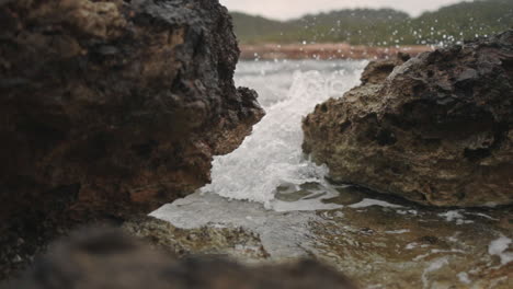 Wave-crashes-through-rocks-at-the-beach,-slow-motion,-low-gimbal-zoom-in