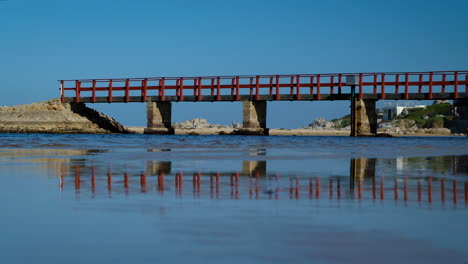Red-bridge-over-lagoon-reflected-in-wet-sand,-Kleinmond,-South-Africa