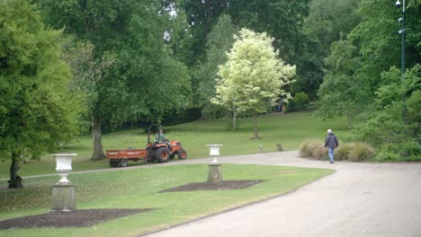 Handheld-panning-shot-of-orange-sit-on-lawnmower-with-trailer-driving-through-Sheffield-Botanical-Gardens-England