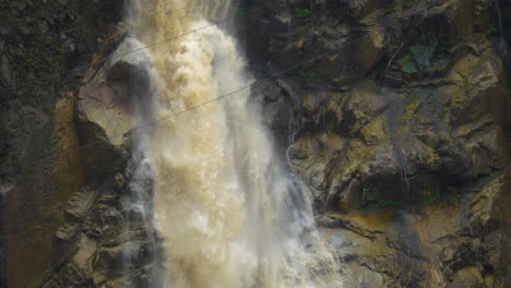 Dirty-water-crashing-down-the-waterfall-in-slow-motion-along-rocky-wet-cliff-wall---Banos,Ecuador