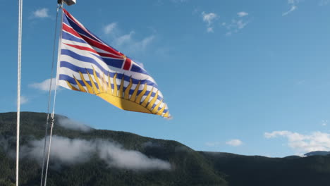 British-Columbia-flag-flutters-against-blue-sky-and-forested-mountain