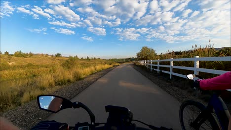 Cyclists-riding-down-a-nature-trail-beside-a-river-with-the-morning-sun-and-sky-reflecting-off-the-water---first-person-view