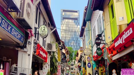 Colorful-Signboards-On-Storefronts-Of-Cafes-At-Haji-Lane-In-Singapore-With-Pan-Pacific-Hotel-In-Background