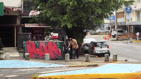 Old-asian-man-sweeps-and-shovels-floor-dirt-into-a-trash-bag-to-put-it-temporary-storage-container-structure-with-graffiti-for-the-garbage-truck-to-pickup-next-to-Spain-Avenue-in-Panama-City