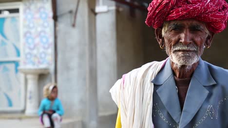Close-up-of-old-senior-Indian-man-wearing-red-turban-leaning-on-yellow-stick-and-child-sitting-on-stairs-in-background