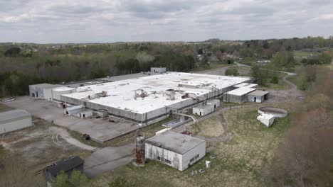 Aerial-Drone-Footage-Moving-Towards-an-Abandoned,-Decaying-Factory,-with-a-Truck-Depot-and-Train-Line