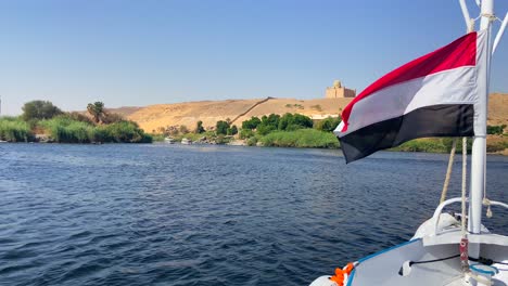 ägyptische-Flagge-Weht-Am-Bug-Eines-Bootes,-Das-Den-Nil-Hinuntersegelt,-Mit-Blick-Auf-Einen-Tempel-In-Der-Wüste