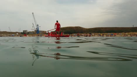 Red-Italian-lifeguard-ready-for-first-aid-emergency-standing-and-rowing-rescue-boat-while-watching-people-swimming-and-beach-in-background-at-Punta-Penna-in-Italy