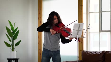 Latino-musician-with-long-hair-in-jeans-and-grey-shirt-playing-song-from-sheet-music-on-red-viola-in-white-studio