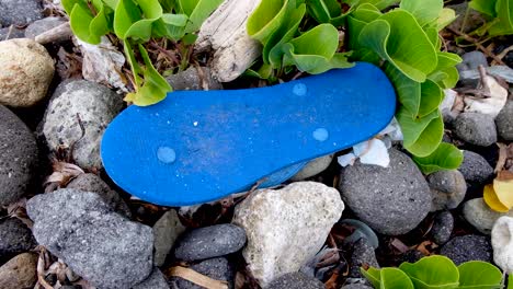 Ocean-and-beach-plastic-and-rubbish-clean-up,-one-of-many-rubber-flip-flop-littering-on-the-pebbles-along-the-shoreline-of-a-popular-tropical-island-destination-in-Southeast-Asia