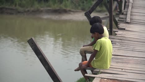 Two-poor-kids-sitting-on-wooden-bridge-on-a-river-in-village,-two-little-friends-at-rural-india-scene