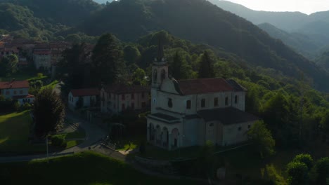 Aerial-View-Of-Scenic-Italian-Village-Parish-Church-Located-Beside-Forested-Hills-In-Arola,-Italy
