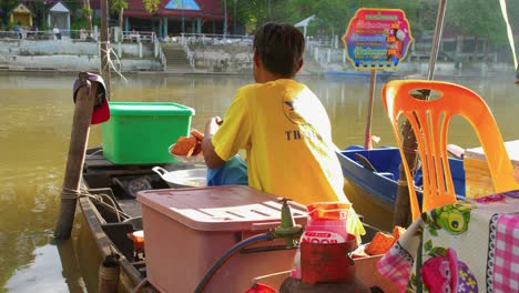 Thailändischer-Mann-Mit-Dem-Rücken-Zur-Kamera,-Der-Auf-Einem-Schwimmenden-Markt-Typisch-Asiatische-Straßengerichte-Am-Ufer-Des-Flusses-Brät