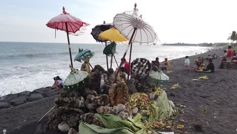 Balinese-Temple-at-Local-Beach,-Purnama,-Bali-Families-Enjoy-Hindu-Umbrellas-and-Deities-Statues-placed-near-the-Waves,-Indonesian-People-in-the-Black-Sand-Spiritual-Ocean,-Sukawati,-Gianyar